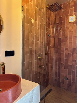 Ein Badezimmer mit einer gläsernen Duschkabine, einem weißen Waschbecken mit roter Schüssel und einer gefliesten Wand.