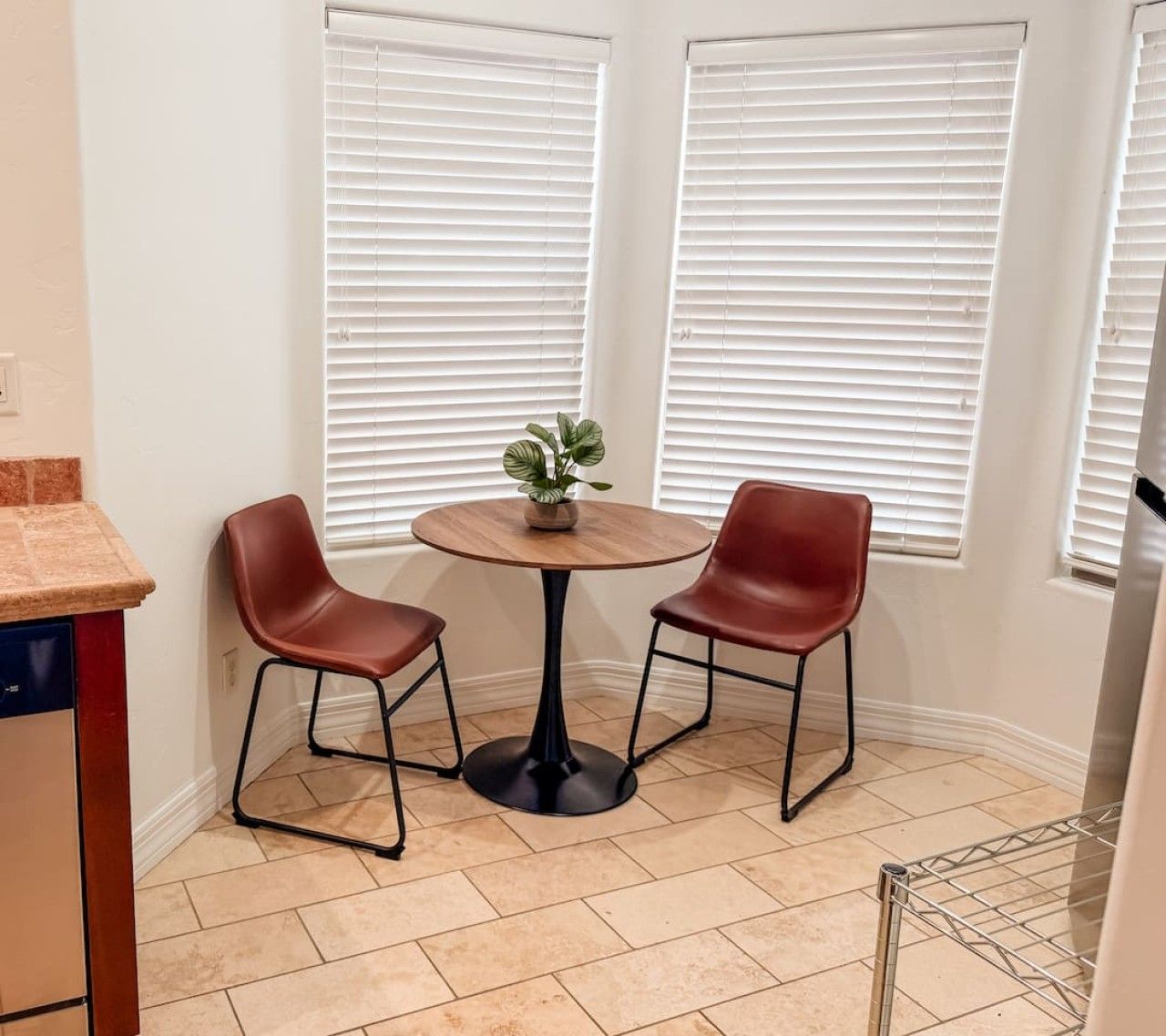 A small dining area with a round table and two chairs, a potted plant on the table, and white blinds on the windows.