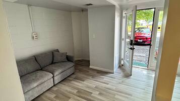 Quarto com sofá cinza, piso de madeira e porta que dá para o exterior.