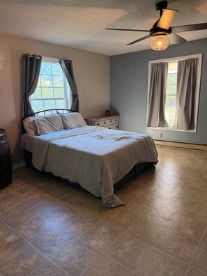 A bedroom with a bed, a dresser, a window with curtains, a ceiling fan, and a tiled floor.