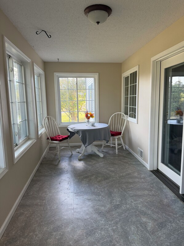 A dining area with a round table, two chairs, a vase with flowers, and a view through a window.