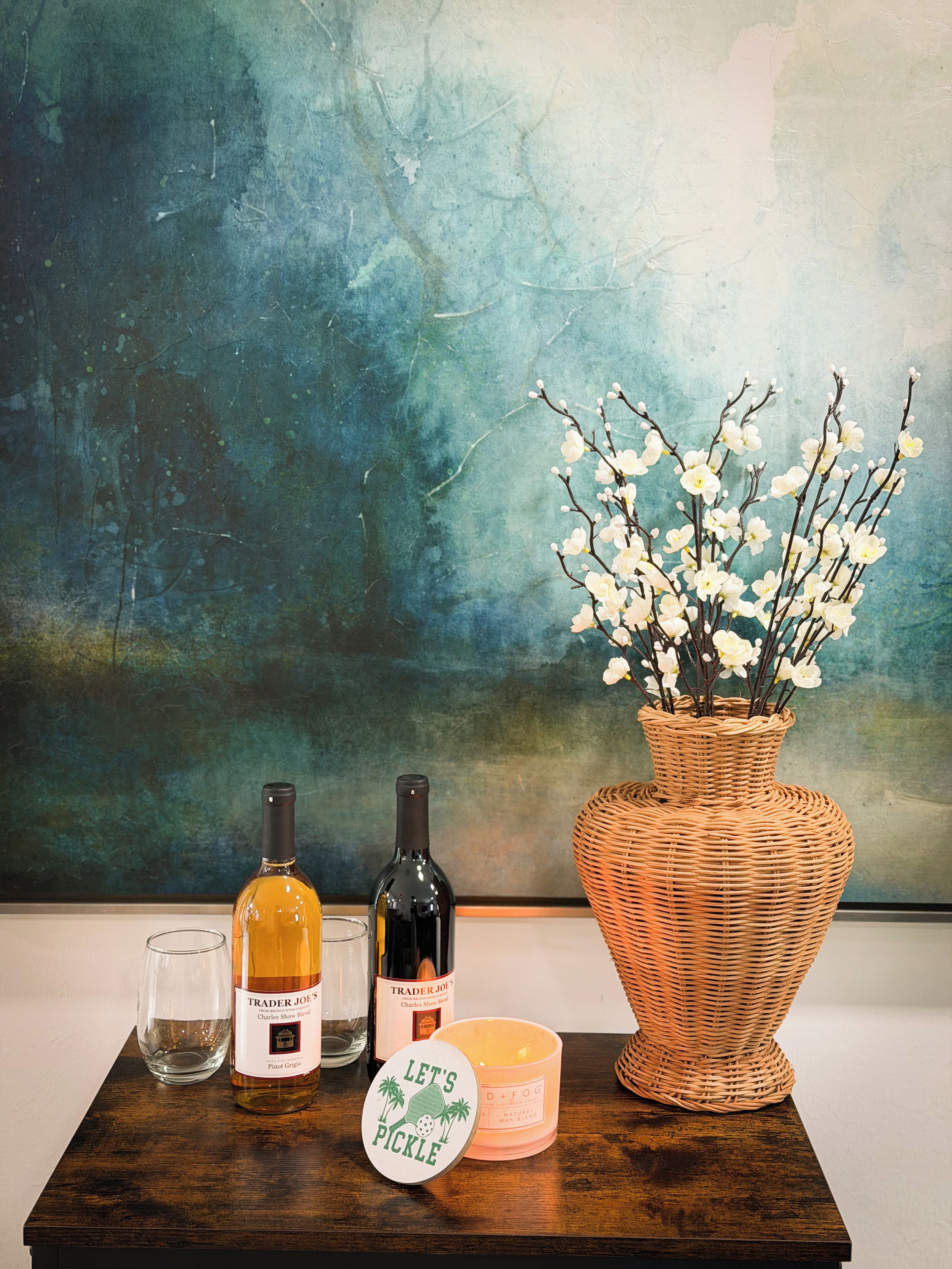 A wooden table with two wine bottles, a candle, and a vase with white flowers against a textured wall.