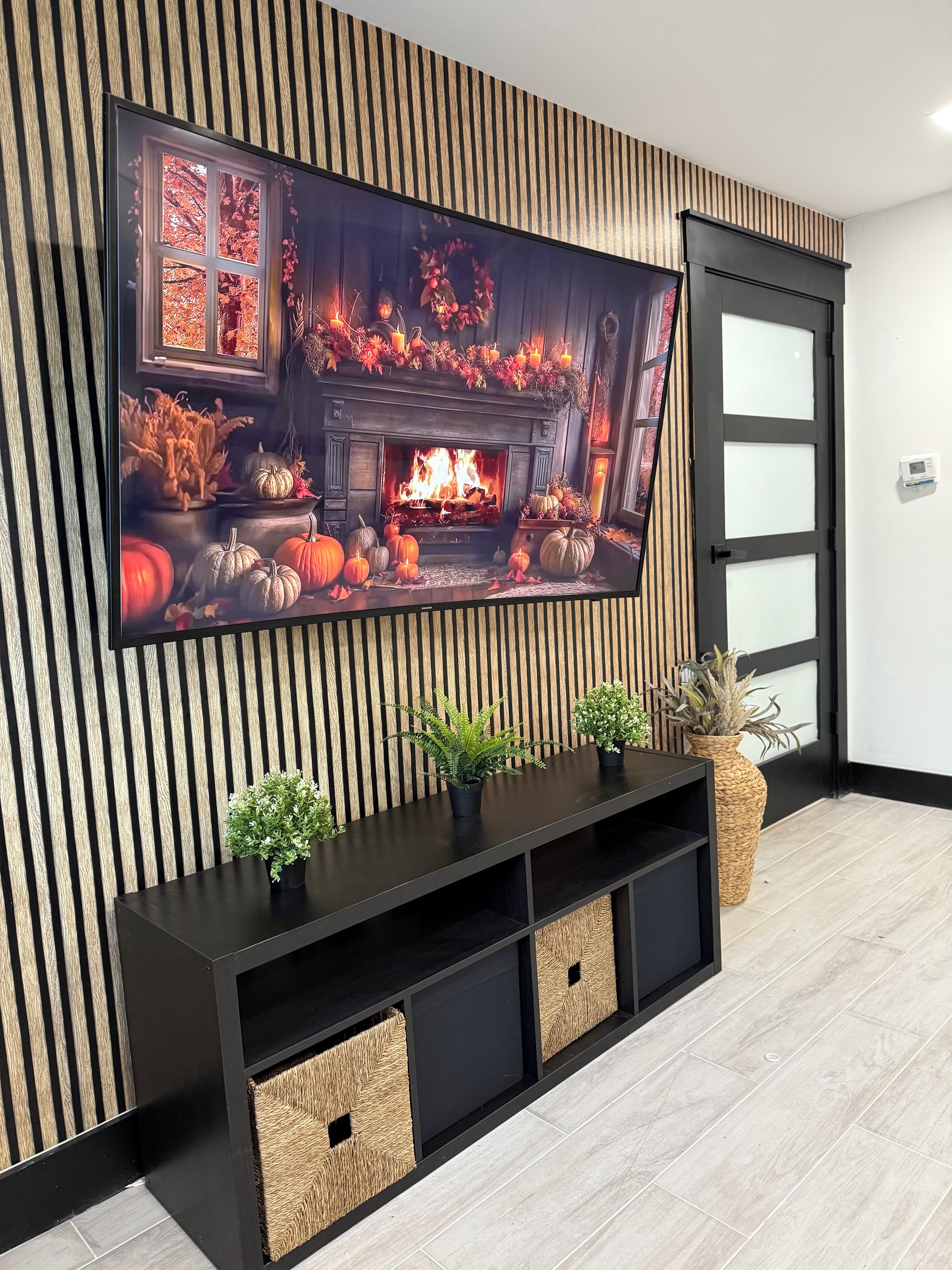 A modern living room with a large flat-screen TV mounted on a wooden wall, a black entertainment unit below, and a fireplace with seasonal decorations.
