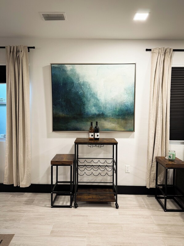 A room with a large abstract painting, a small wooden table with wine bottles, and a window with curtains. - Near Downtown Pickleball Paradise (Naples)