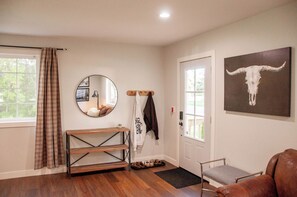 A room with a wooden bench, a coat rack with a jacket, a mirror, a door with a bull skull painting, and a window with curtains. - Charming Crookston Cottage (Crookston)