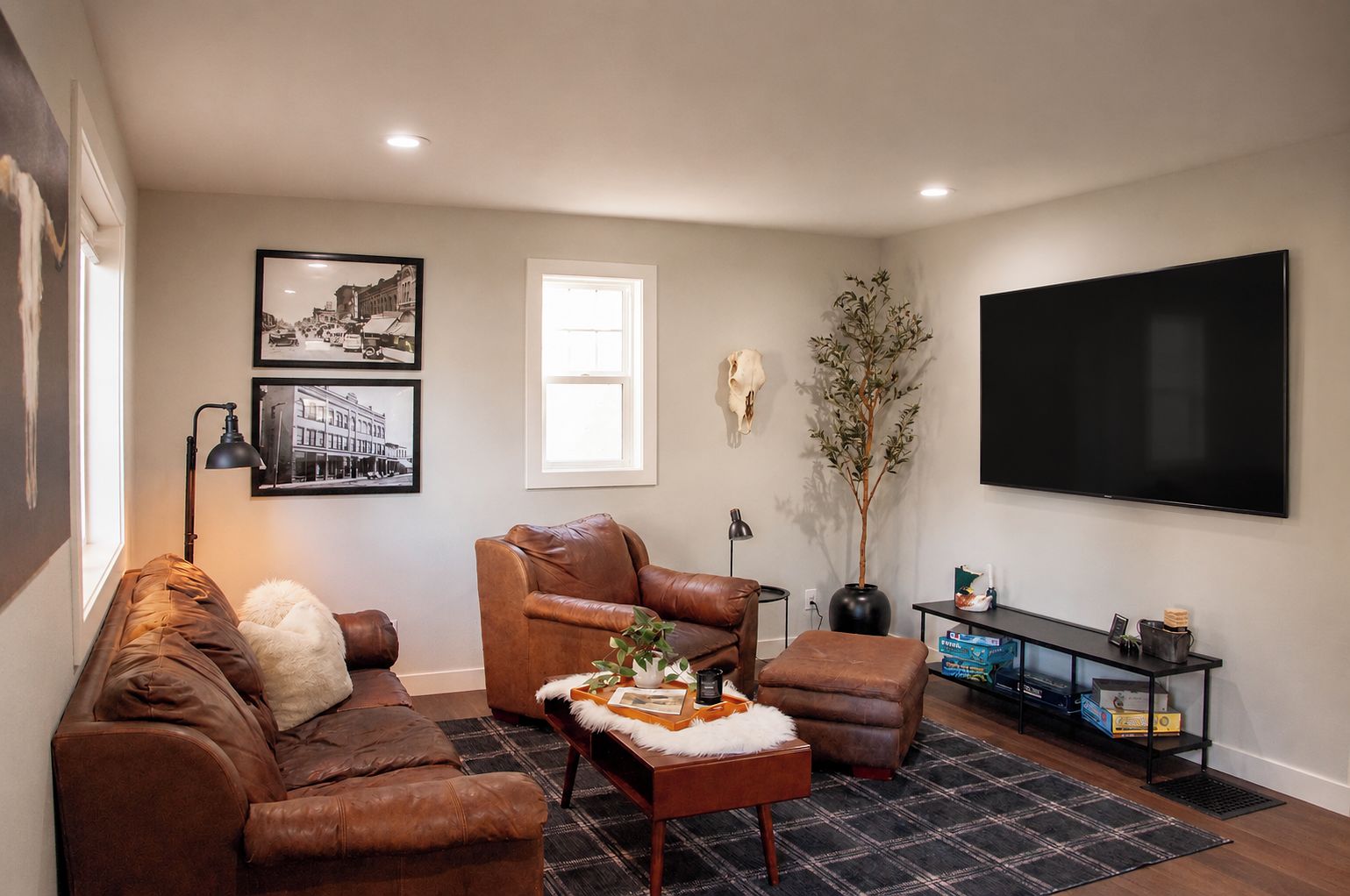 Ein Wohnzimmer mit braunem Ledersofa, einem braunen Ledersessel, einem Couchtisch, einem Flachbildfernseher, einem gerahmten Bild, einer Vase mit Pflanze und einem kleinen Tisch mit Lampe.