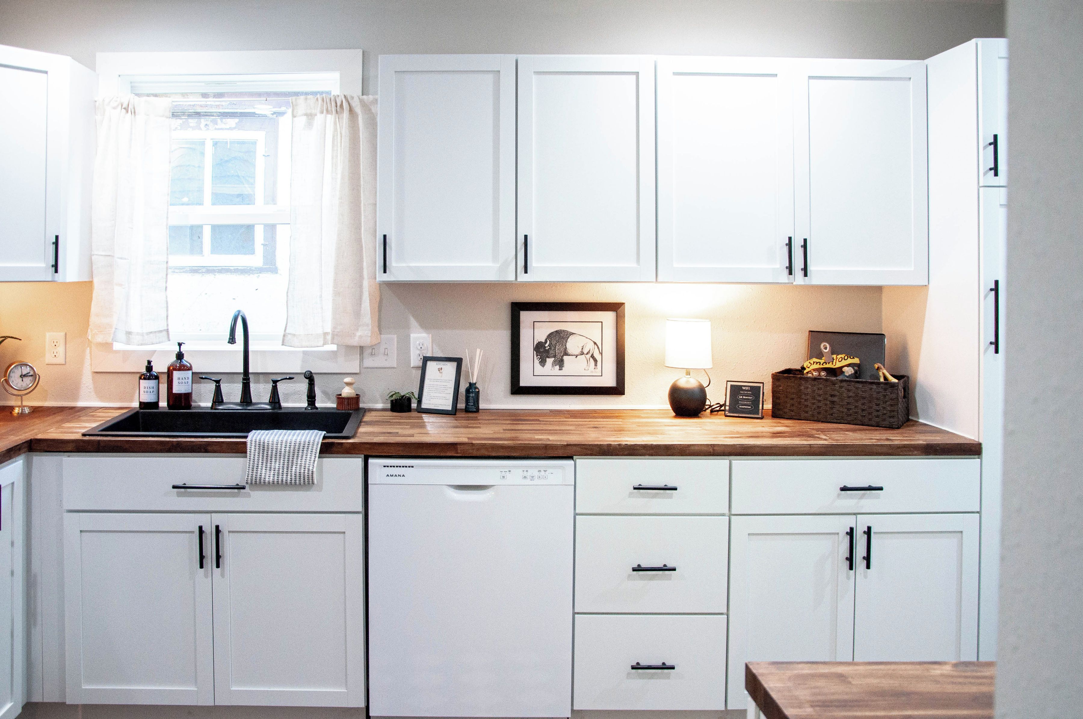 A kitchen with white cabinets, a wooden countertop, a dishwasher, and a window with curtains.