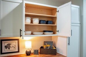 A kitchen with open cabinets displaying dishes and bowls, a framed picture on the counter, a lamp, and a basket with items.