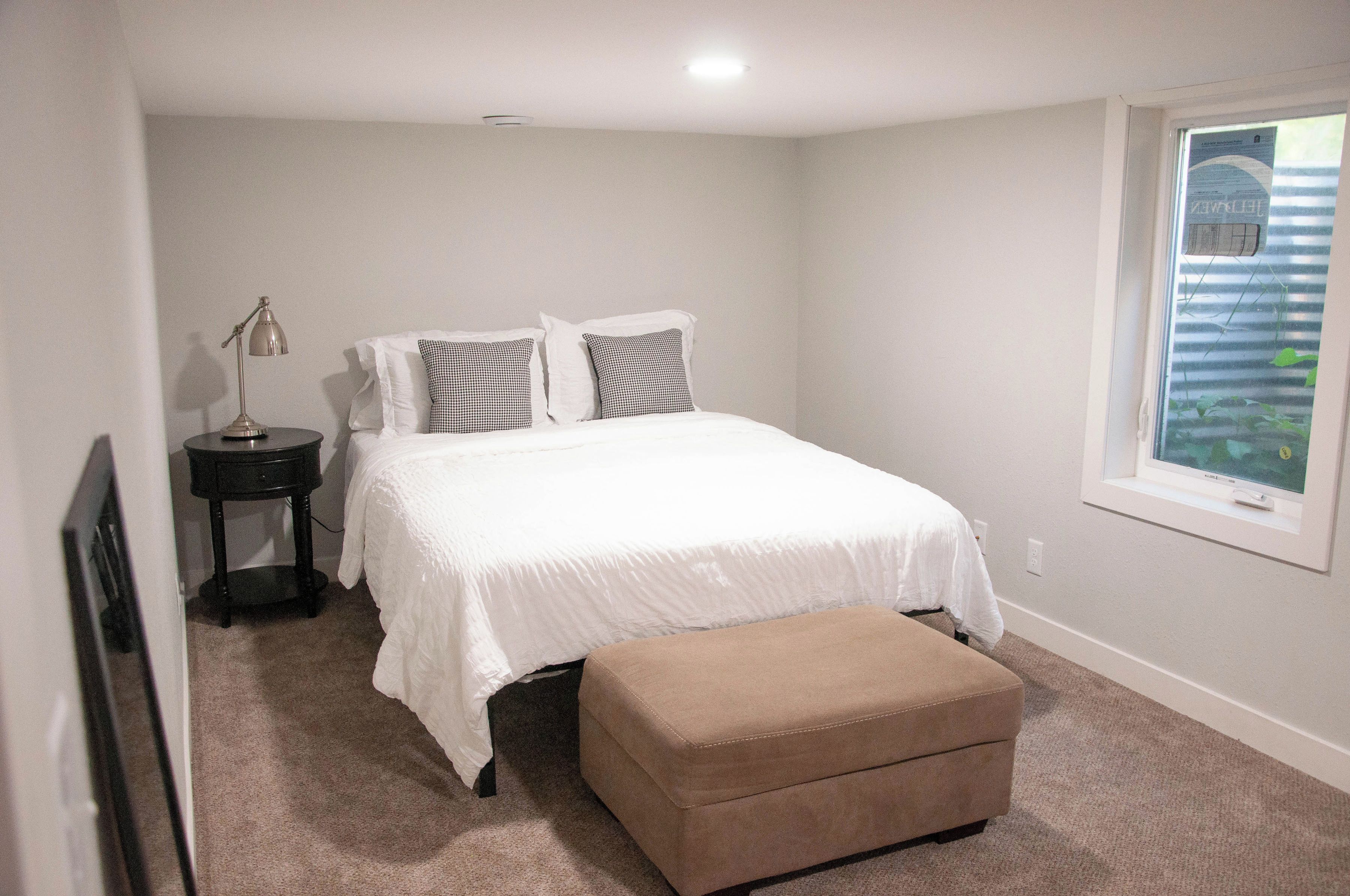 A bedroom with a bed, a nightstand, a lamp, a window, and an ottoman.