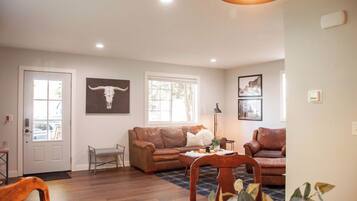 A living room with a brown leather sofa, a small bench, and a dining table with chairs. There are two windows, a door, and a ceiling light fixture.