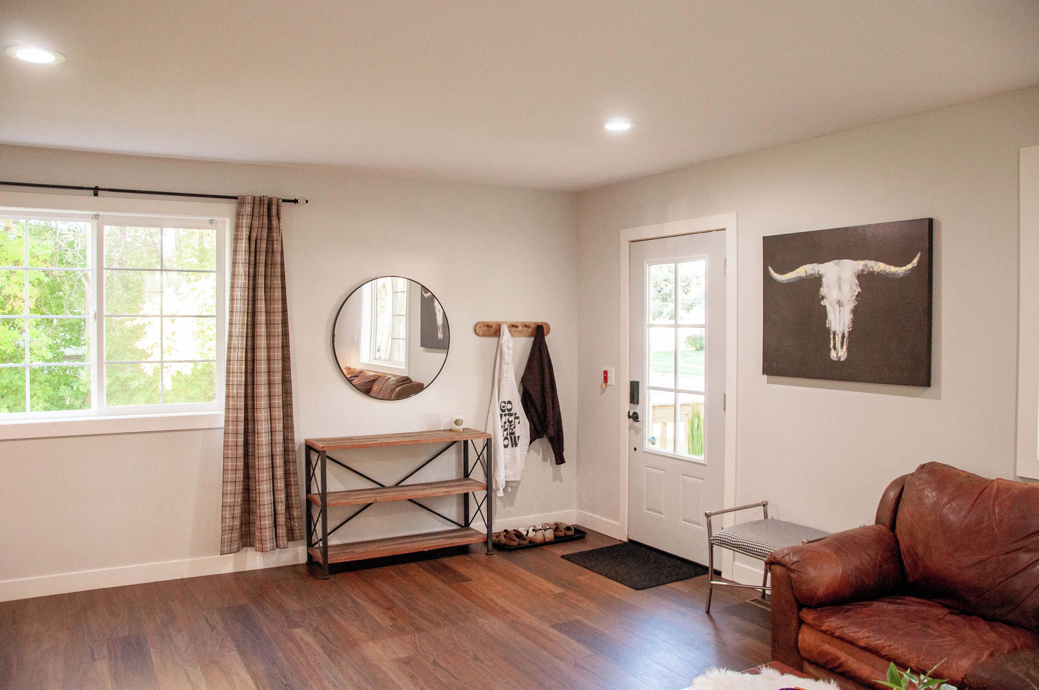 A room with a wooden floor, a brown leather couch, a door with a mirror, a wall-mounted artwork of a bull's head, and a window with a plaid curtain.