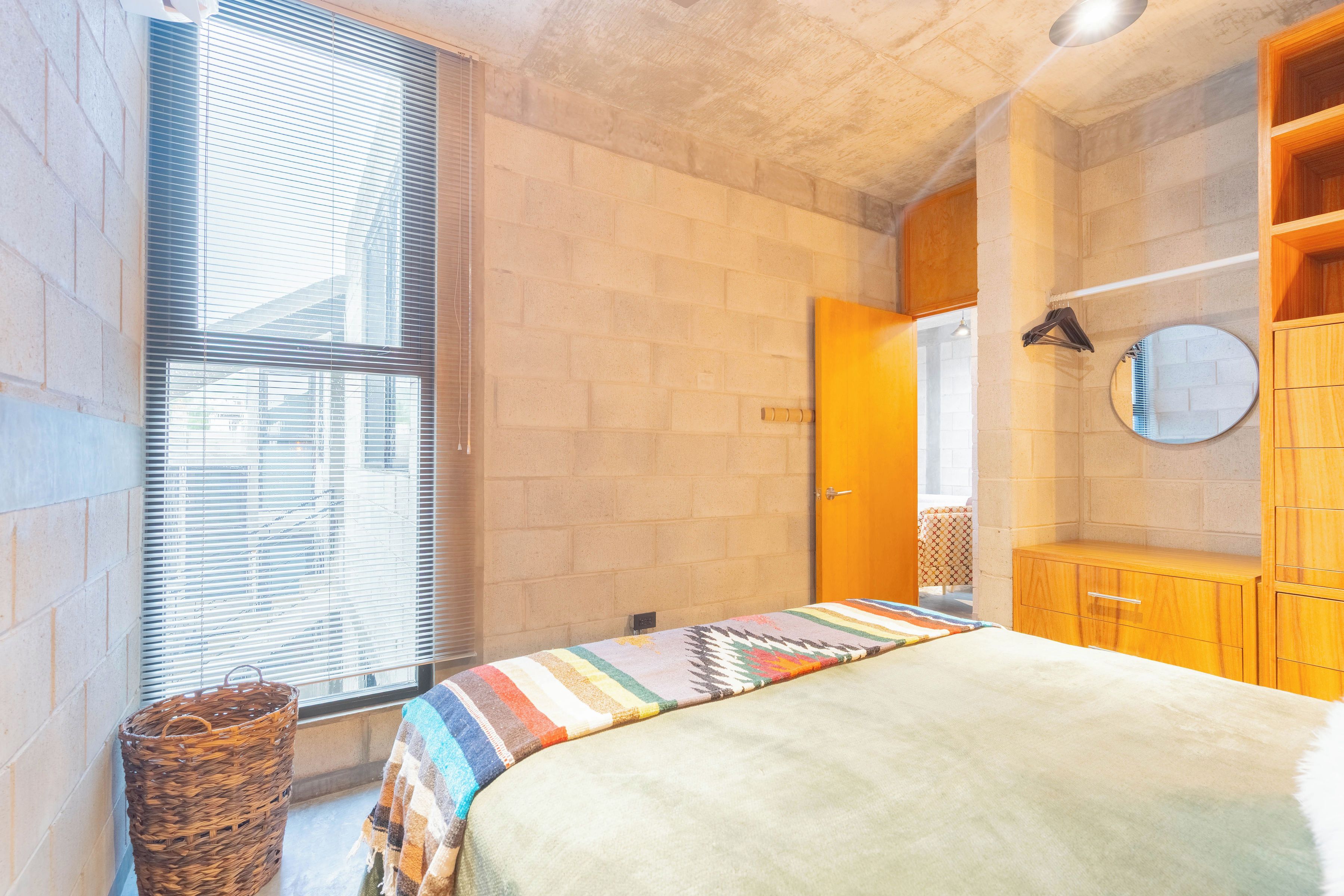 Ein Zimmer mit einem Bett, einem Fenster mit Jalousien, einem Holzschrank und einem Spiegel an der Wand.