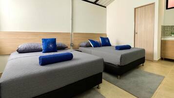 A hotel room with two beds, each with a grey mattress and blue pillows. There is a wooden headboard, a grey rug, and a wooden door.