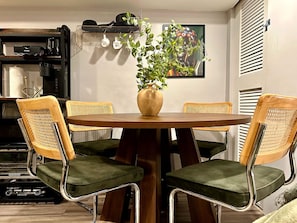 A dining area with a round table, four chairs, a vase with a plant, a coffee machine, and a shelf with kitchenware.