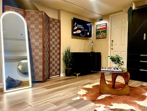 A modern living room with a wooden floor, a large mirror, a flat-screen TV, a potted plant, and a unique wooden coffee table.