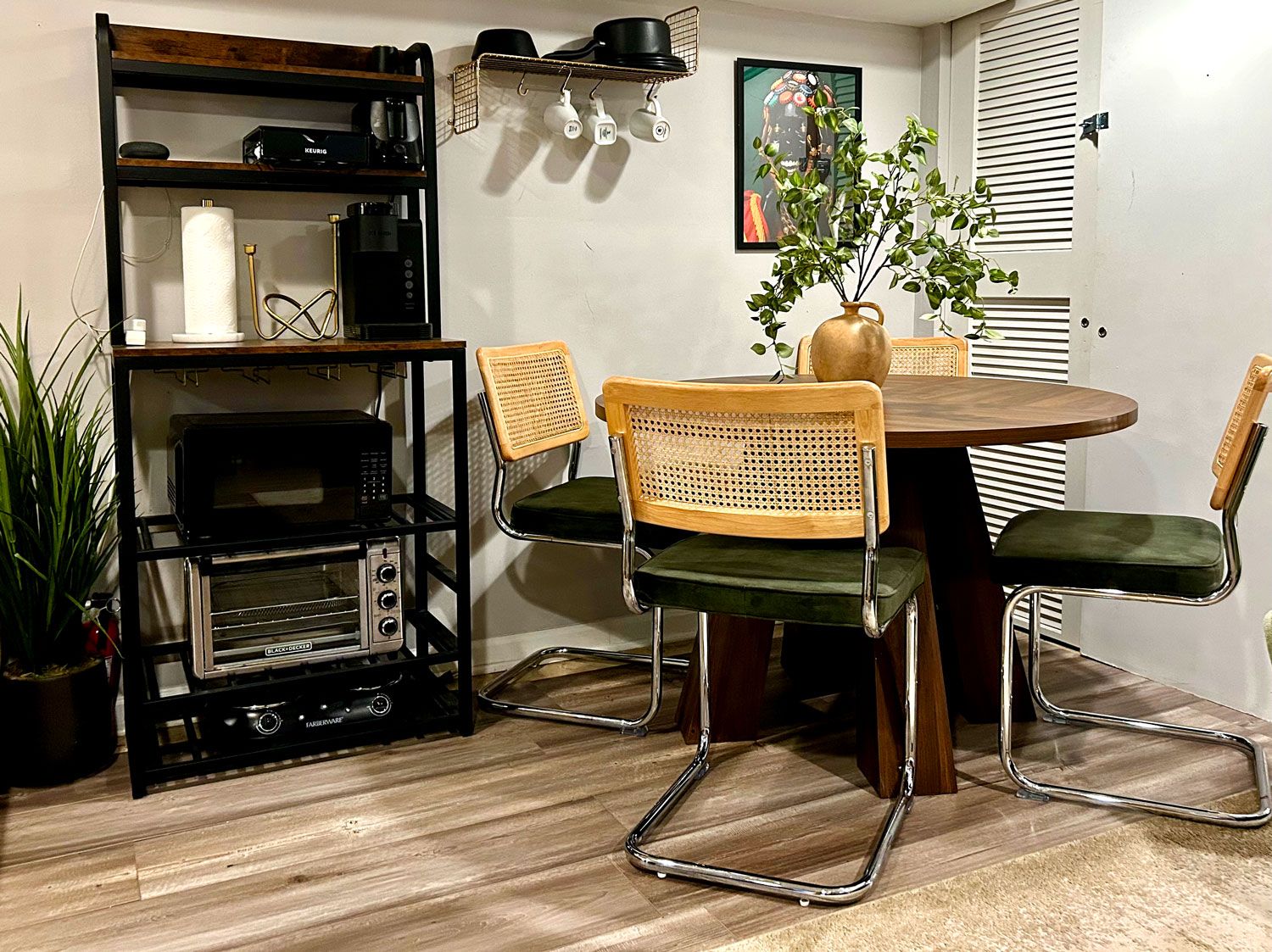 A dining area with a round table, two chairs, a potted plant, a shelf with kitchen appliances, and a wall-mounted coffee machine.