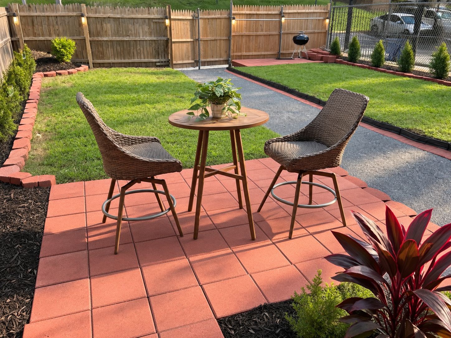A patio with two wicker chairs and a small round table, surrounded by a brick border and a grassy lawn.