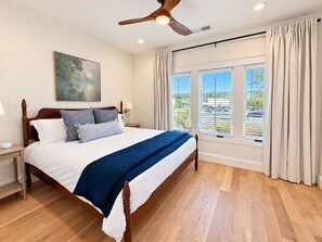 A bedroom with a large bed, a ceiling fan, a painting, a nightstand, a lamp, and a window with curtains. - Homewood Luxury 2 Bedroom 2 Bath Townhome 3020 (Homewood)