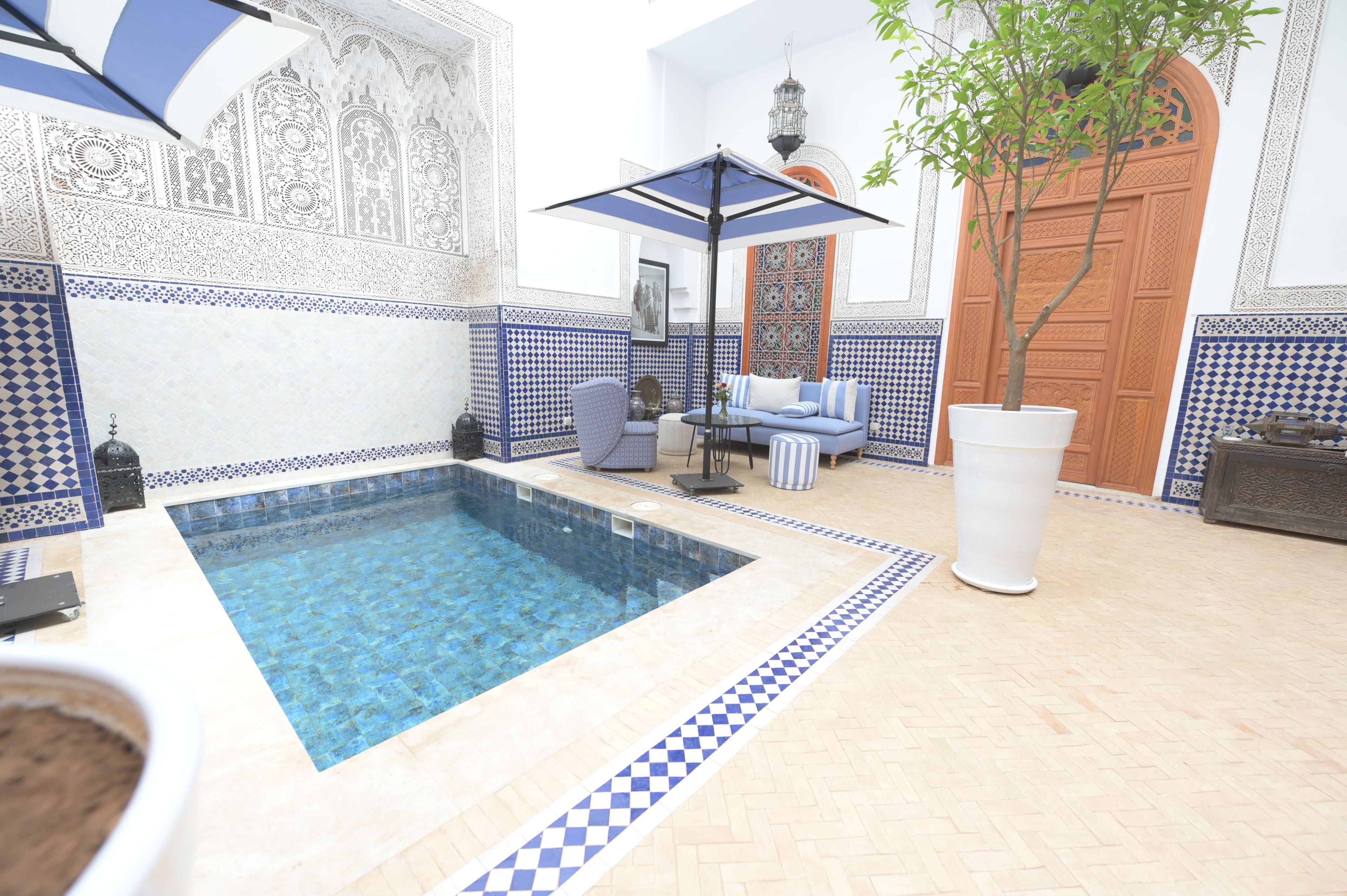 Zona de piscina con suelo de baldosas azules y blancas, una sala de estar con sofá y mesa, y una pared decorativa con diseños intrincados.