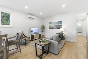 A modern living room with a flat-screen TV, a grey sofa, a wooden dining table with chairs, and a small coffee table. - Carriage on Richter •2 King Beds• Sleeps 6 • Ski Base • Close to Hospital (Kelowna)