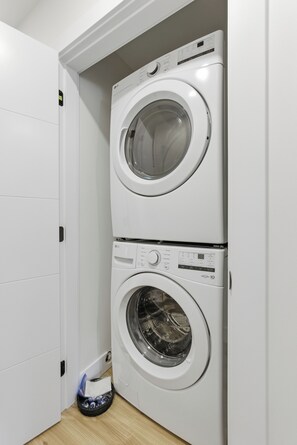 A compact laundry area with two stacked washing machines, a basket with laundry items, and a wooden floor. - Carriage on Richter •2 King Beds• Sleeps 6 • Ski Base • Close to Hospital (Kelowna)