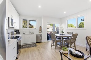 A modern kitchen with white cabinets, a dishwasher, and a dining area with a wooden table and chairs. - Carriage on Richter •2 King Beds• Sleeps 6 • Ski Base • Close to Hospital (Kelowna)