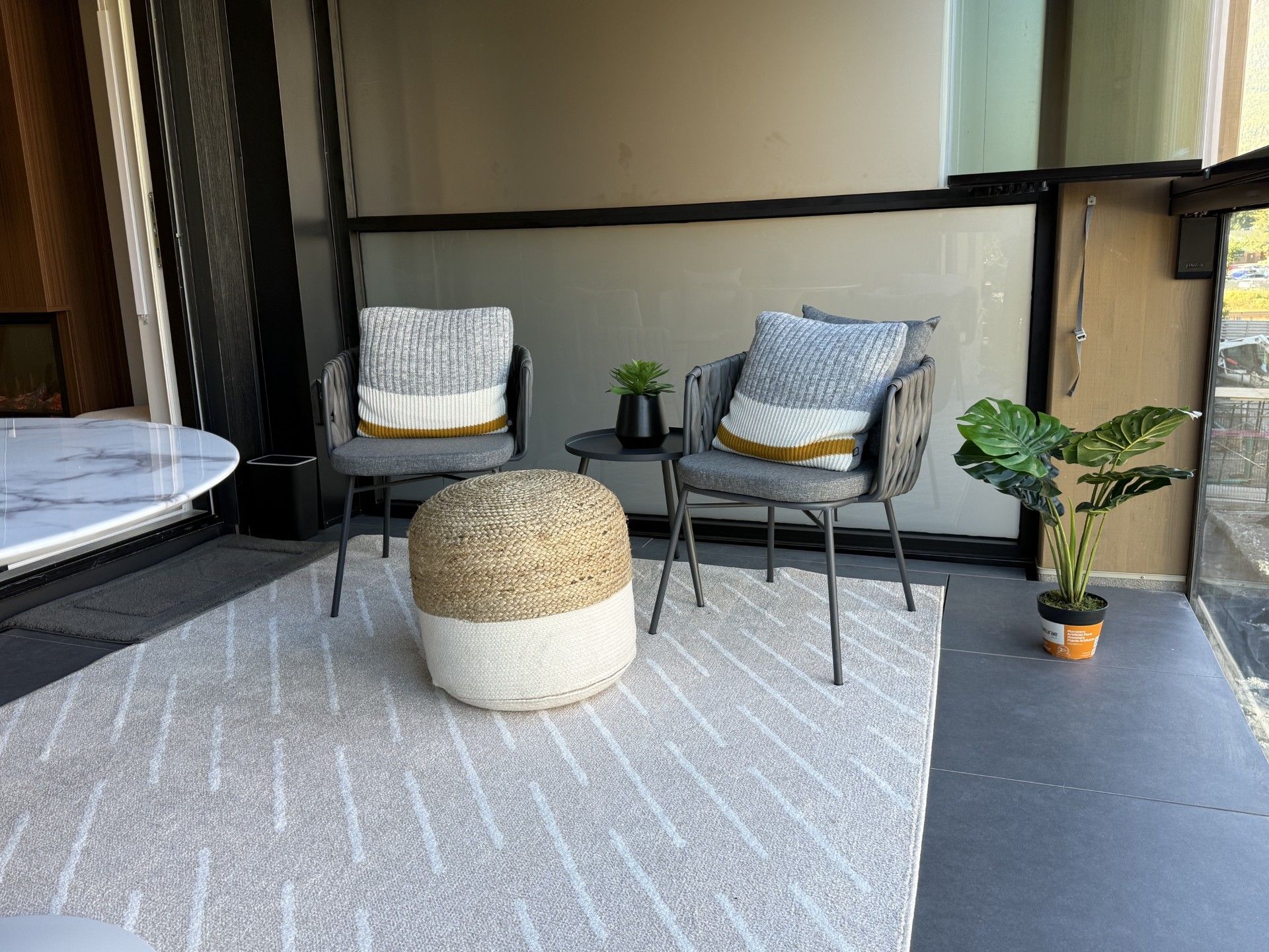 A modern outdoor seating area with a round table, two chairs, a woven ottoman, and potted plants.