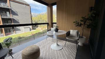 A balcony with a round table, two chairs, a potted plant, and a woven pouf.