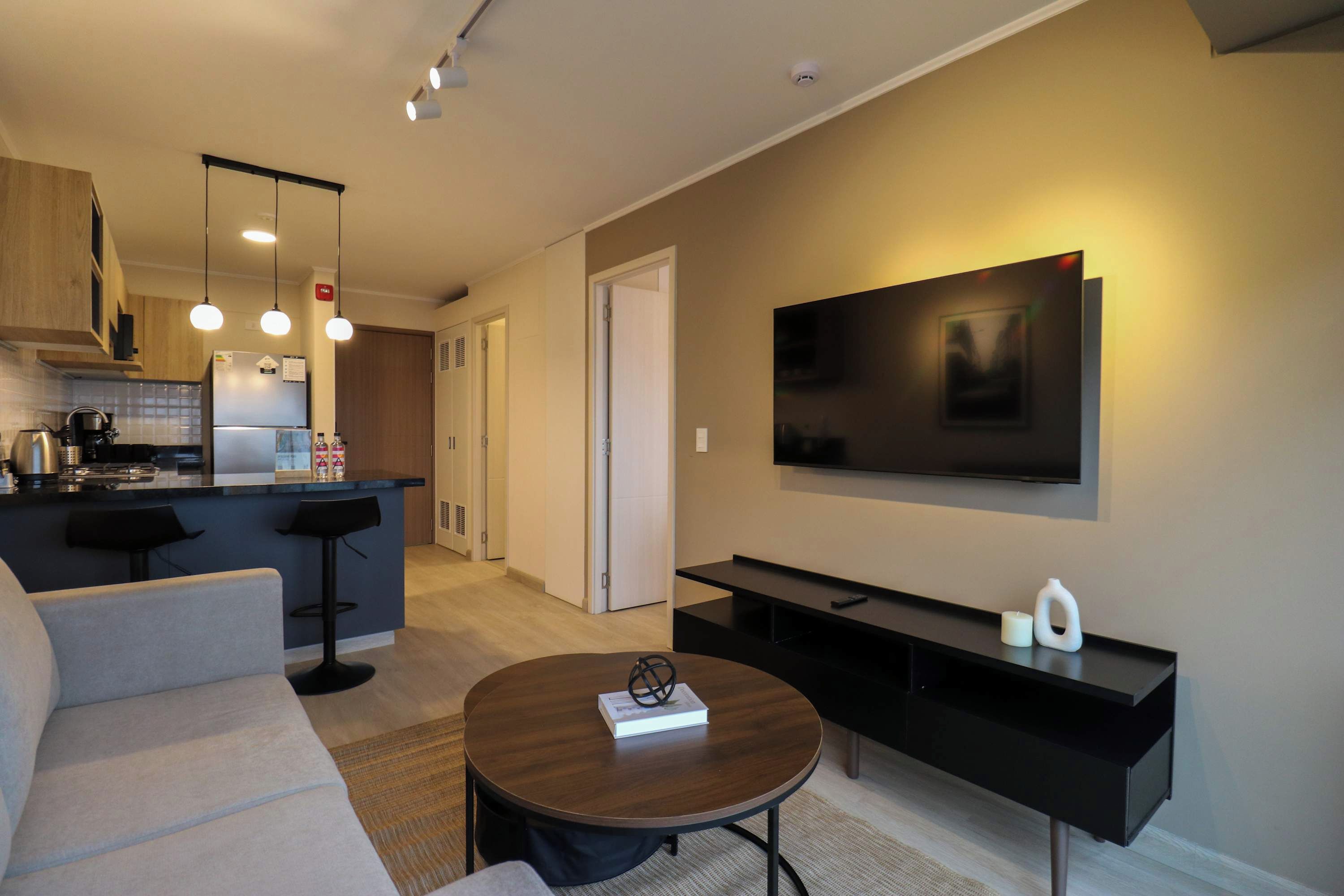 Una sala de estar moderna con un sofá, una mesa de centro, un televisor y una zona de cocina al fondo.