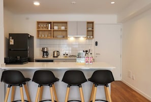 A modern kitchen with a white countertop, wooden bar stools, a refrigerator, and a coffee maker. - w* | Amazing 2BR w/ Partial Sea View in Miraflores (Miraflores)