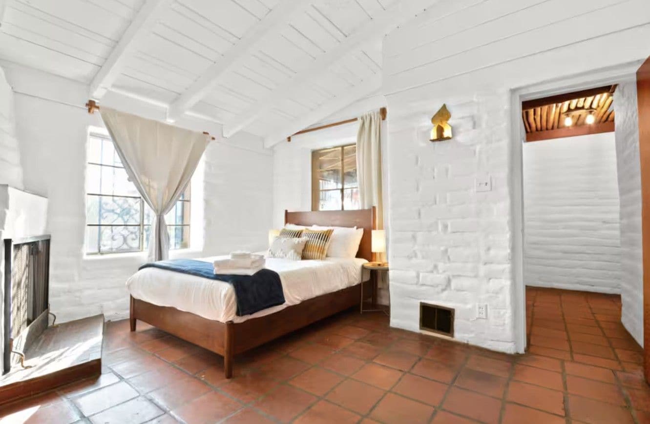 A bedroom with a wooden bed, a nightstand, a window with curtains, and a white brick wall.