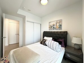 A bedroom with a bed, a nightstand, a lamp, a painting, and a door. - The Modern Retreat | Luxury 2 Bed, 2 Bath Condominium (Mississauga)