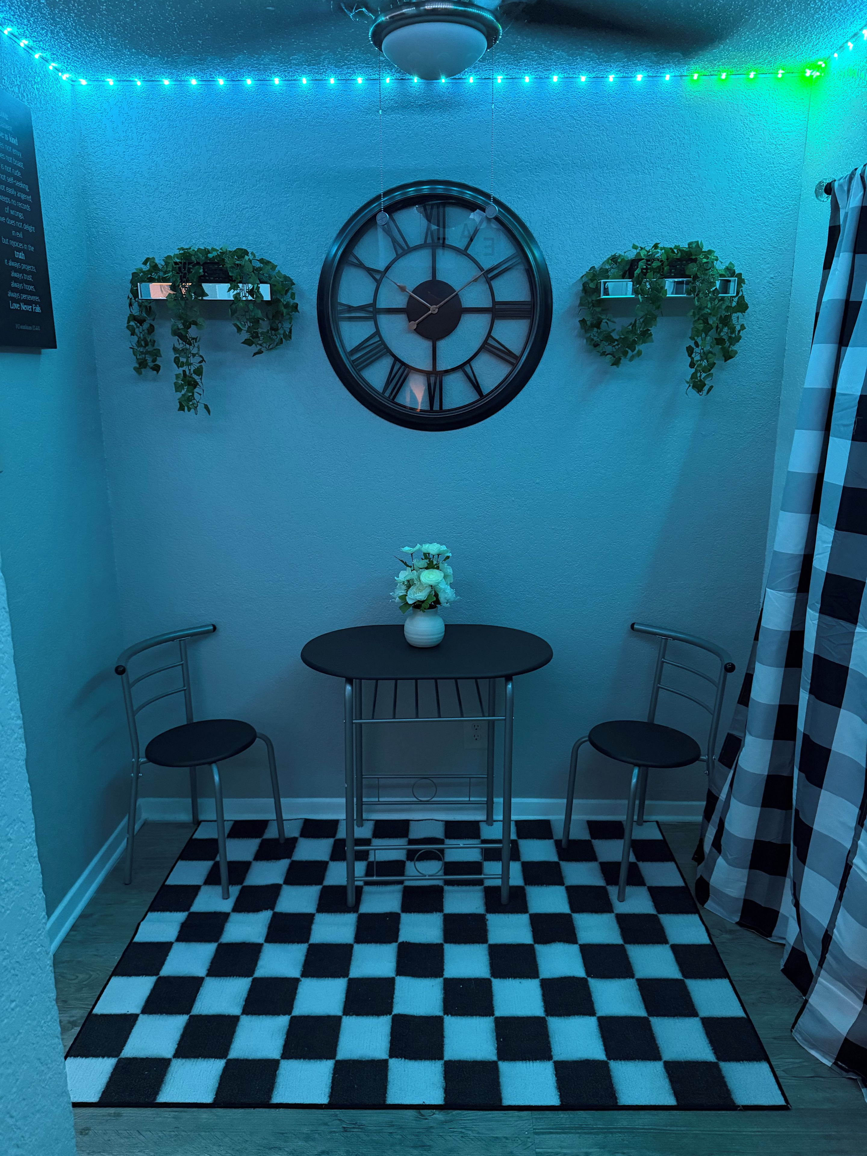 A room with a checkered floor, two chairs, a table with a vase, a wall clock, and green plants.