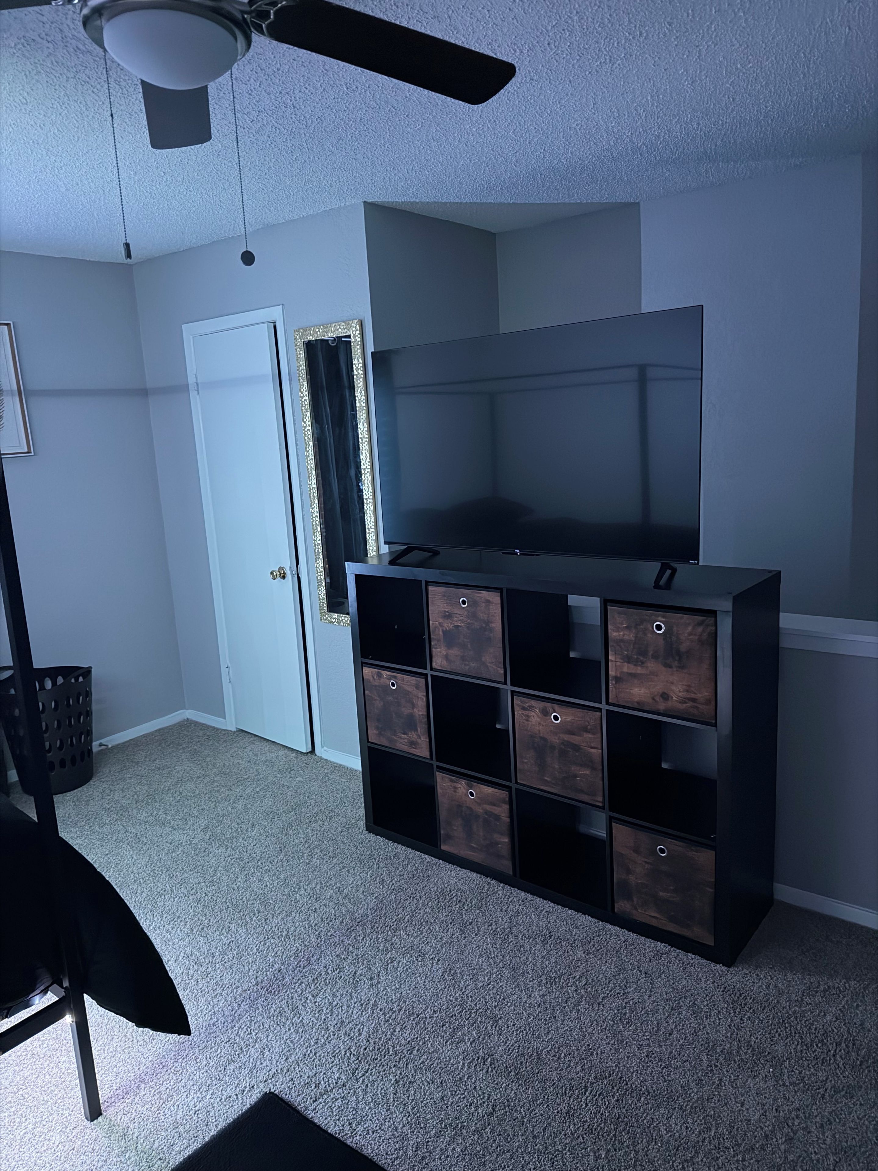 A room with a ceiling fan, a large flat-screen TV, a dark wooden storage unit, a door, and a framed mirror.