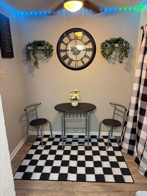 Una habitación con un mantel de cuadros blancos y negros, una mesa redonda con sillas, un reloj de pared y dos estanterías con plantas.