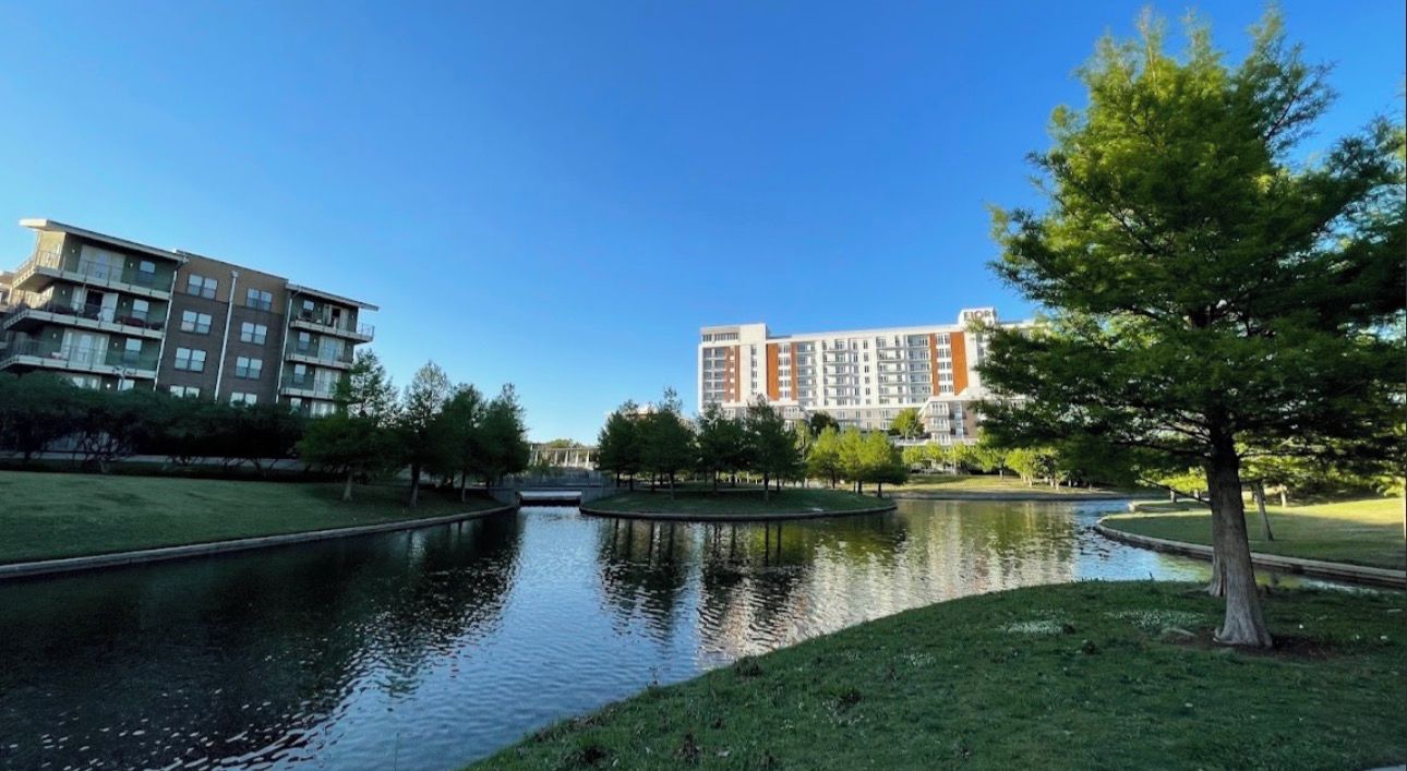 A riverside view with modern buildings, a grassy area, and a small bridge.
