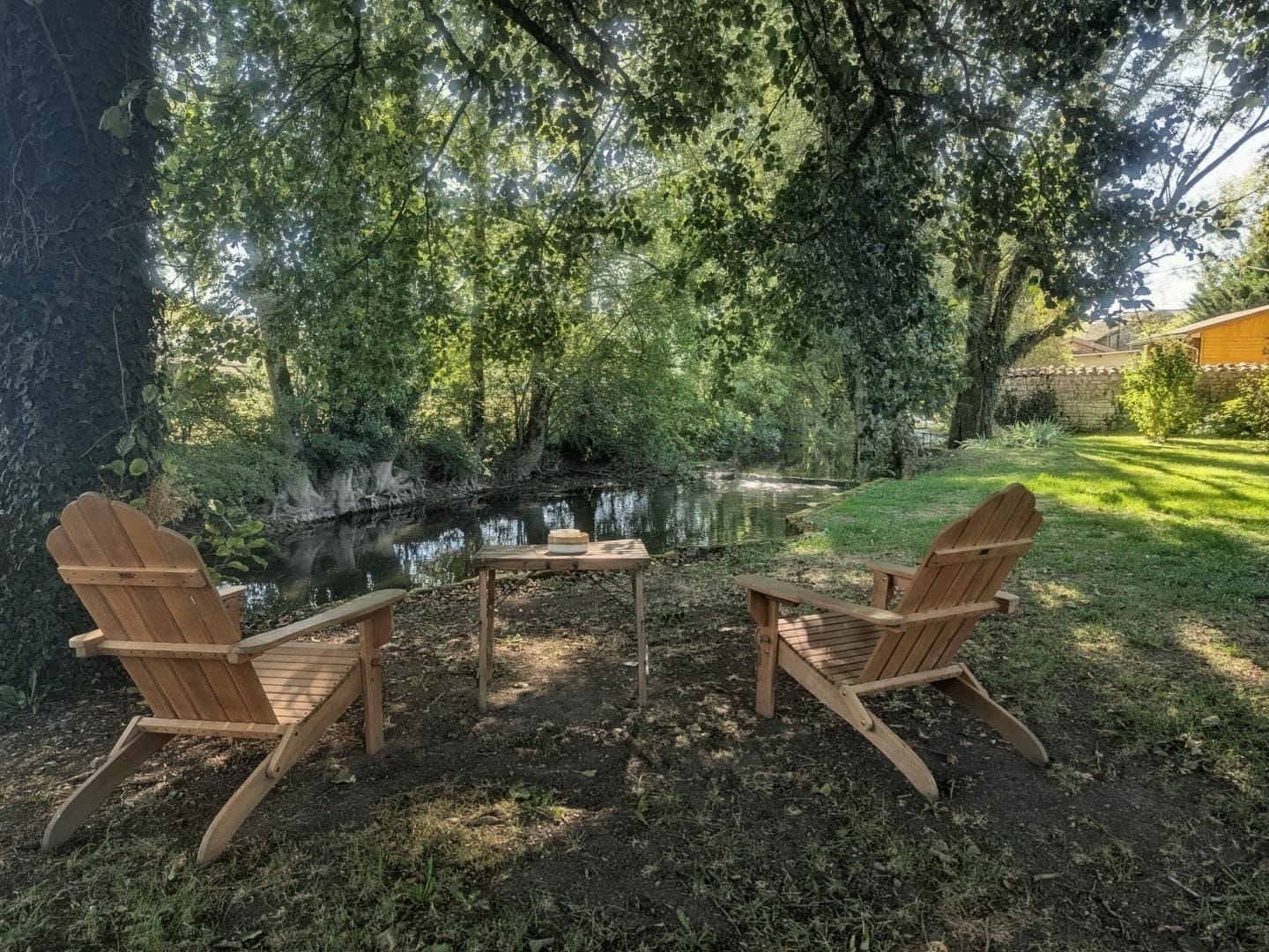 Two wooden Adirondack chairs facing a small table with a box on it, set on a grassy area with a pond and trees in the background.