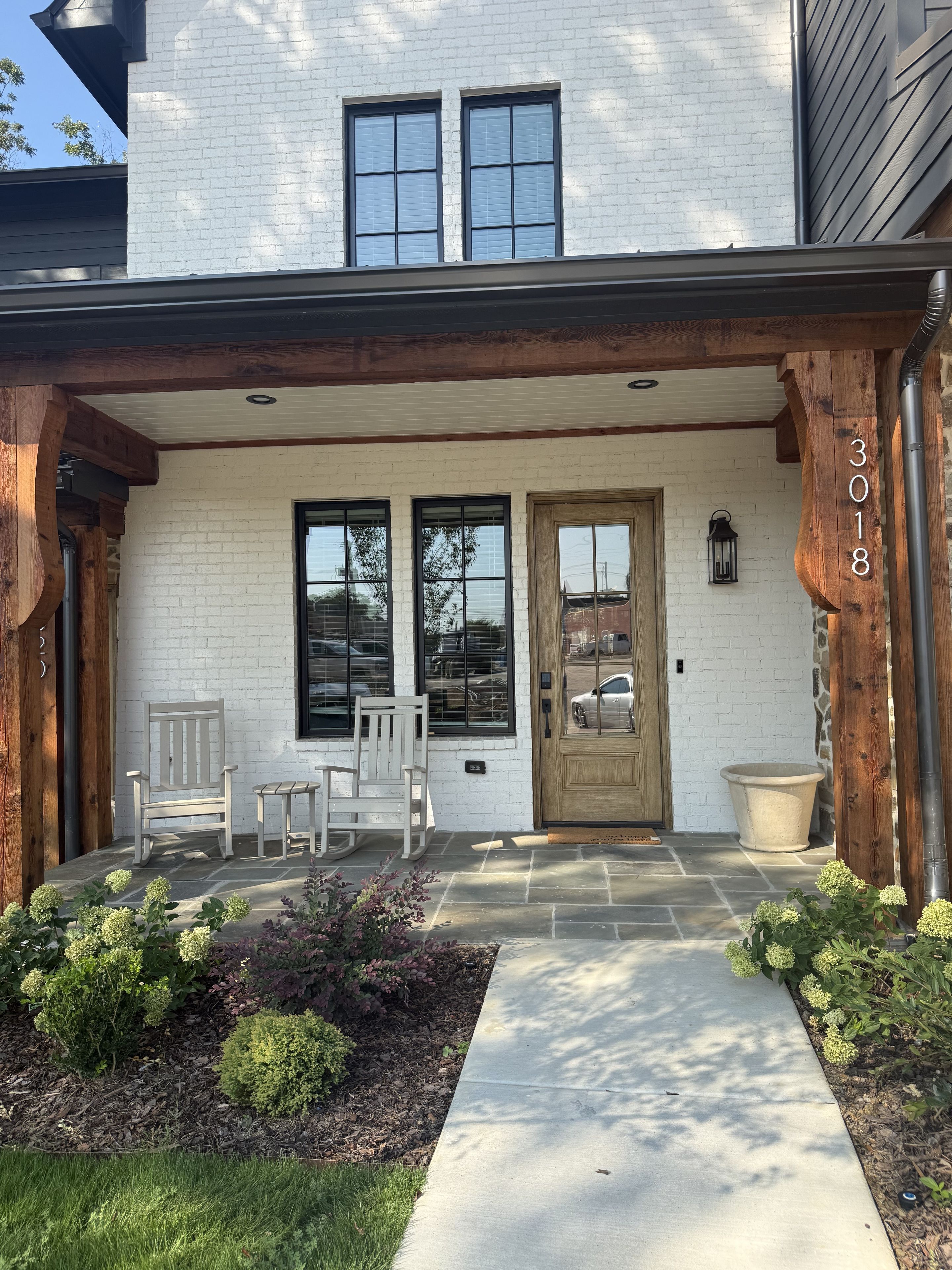 A two-story house with a white brick facade, a front porch with wooden posts, a front door, and two windows.