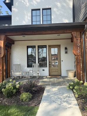 A two-story house with a white brick facade, a front porch with wooden posts, a front door, and two windows.