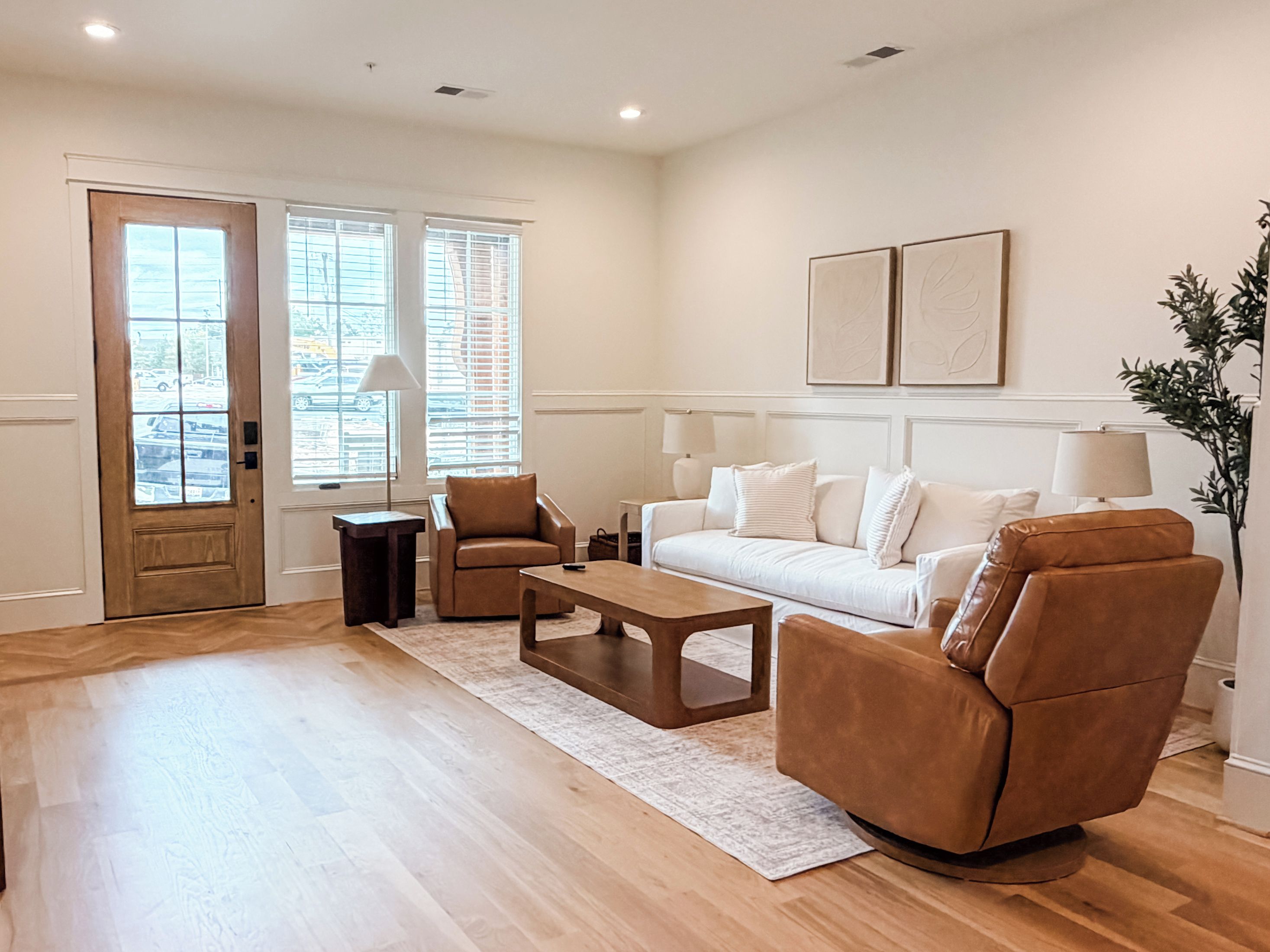 Un salon comprenant un canapé, deux fauteuils, une table basse et une table d’appoint. Il y a deux fenêtres avec des stores, une porte et une plante.