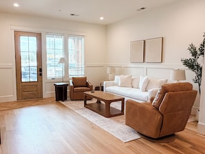 Un salon comprenant un canapé, deux fauteuils, une table basse et une table d’appoint. Il y a deux fenêtres avec des stores, une porte et une plante.
