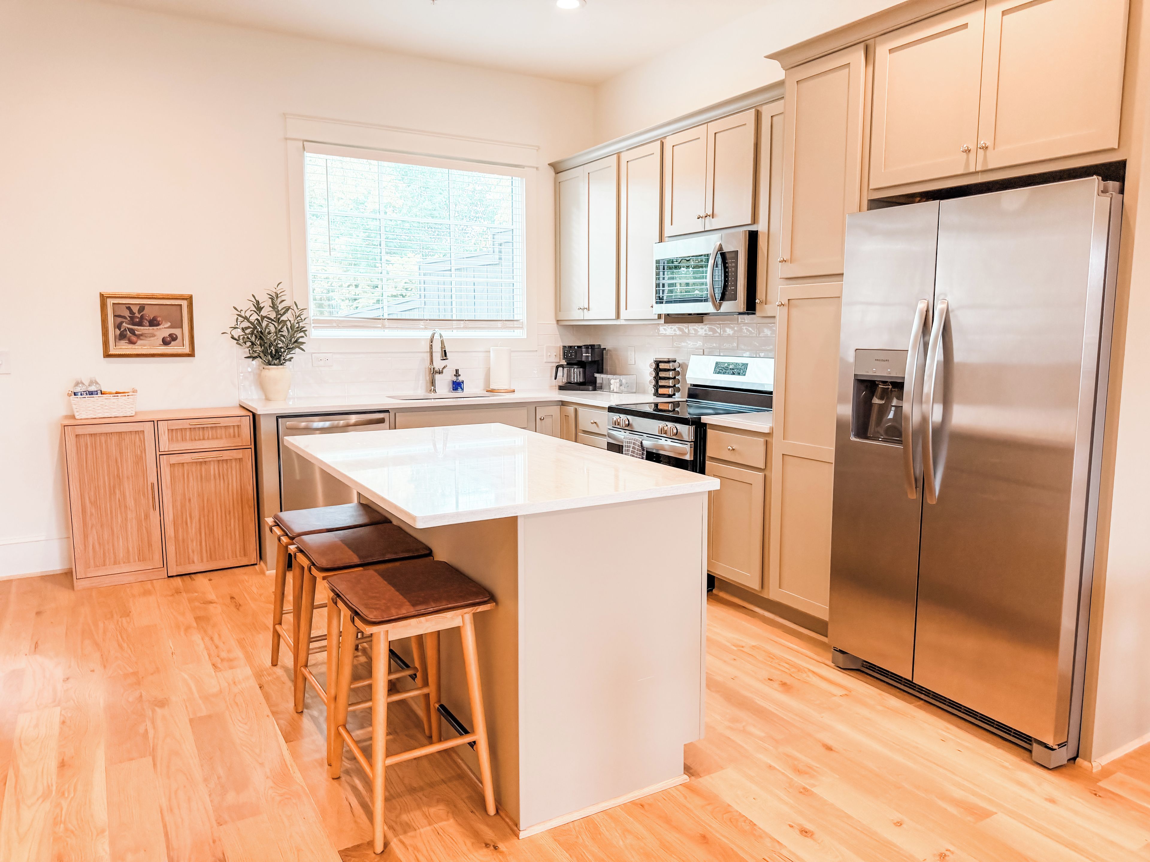 A modern kitchen with wooden cabinets, a stainless steel refrigerator, a central island with stools, and a window providing natural light.