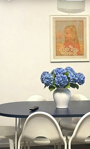 A dining area with a round table, white chairs, a vase of blue flowers, and a framed painting on the wall. - A 13-minute bike ride from downtown Forte dei Marmi: apartment with a terrace (Ripa- Pozzi-Ponterosso)