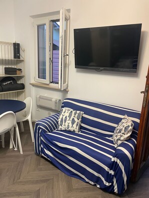 Ein Zimmer mit einer Couch, einem Fernseher, einem Fenster, einem Tisch und Stühlen.