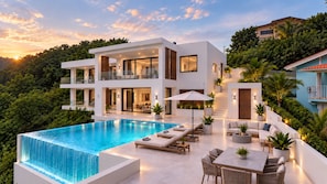 A modern two-story house with a swimming pool, outdoor seating, and lush greenery.