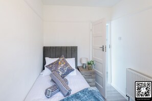 Un letto rifatto con cuscini e coperta, un comodino con una lampada e un vaso, e una porta che dà su un'altra stanza.