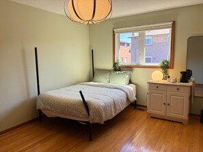 Een slaapkamer met een bed, nachtkastje, raam en spiegel.