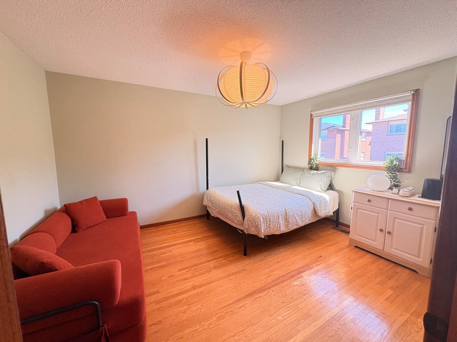 A bedroom with a bed, a red sofa, a bedside table, and a window with a view of buildings.