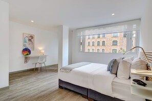 A modern bedroom with a large bed, a window with a view of a brick building, a desk with a chair, and a wall-mounted lamp. - w* | Amazing 2BR in Zona T (Bogotá)