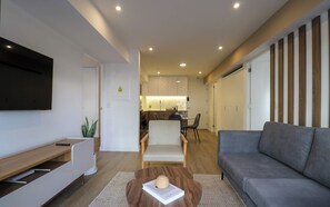 A modern living room with a grey sofa, a coffee table with a book, a TV mounted on the wall, and a kitchen in the background. - w* | Deluxe 2BR w/ Terrace in Miraflores (Miraflores)